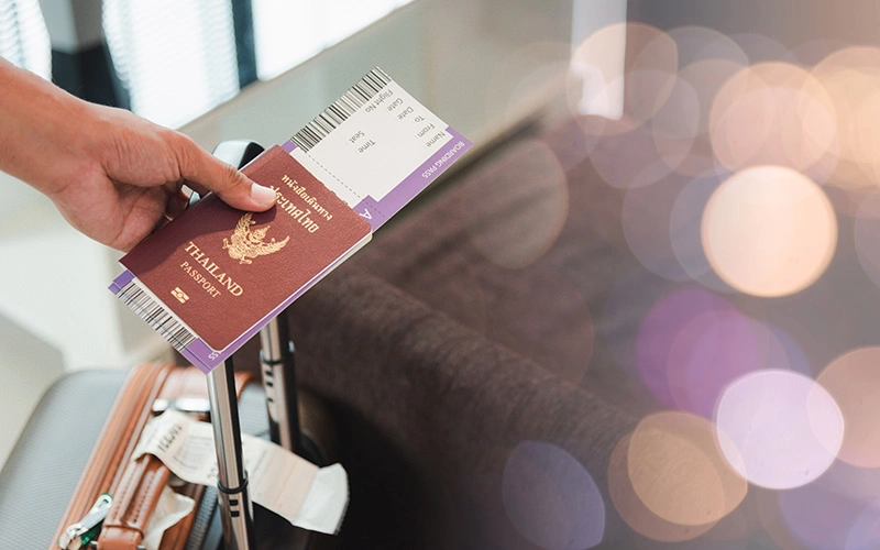Close-up of a man’s hands holding a suitcase and Thailand passport before flight.